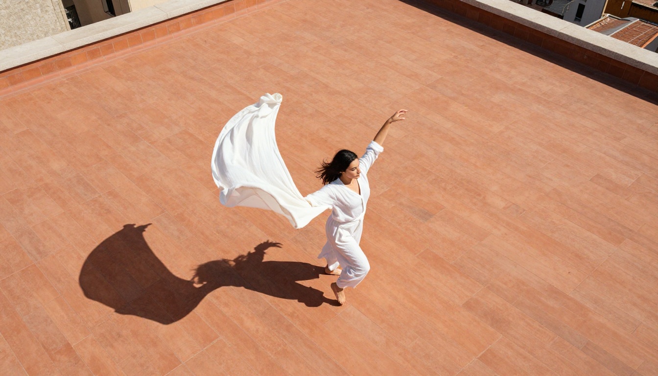 Aerial top-down view of dancer in white linen mid-spin on terracotta rooftop with elongated morning shadows